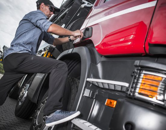 Person in a checkered shirt and cap climbing into a large red truck, with their foot on the step and hand on the door handle.