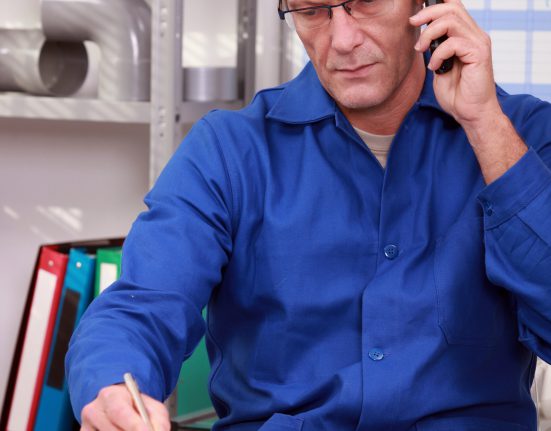 Man in blue shirt talks on phone while writing in a notebook at a desk. Office shelves and a calendar are in the background.