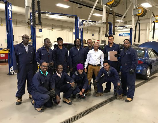 A group of people in blue coveralls and one person in a white shirt pose together in an automotive workshop with cars and equipment in the background.