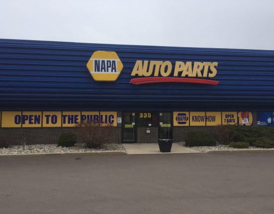 Front view of a NAPA Auto Parts store with blue siding, yellow and white signage, and a banner stating OPEN TO THE PUBLIC. Entrance door at center with pavement in foreground.
