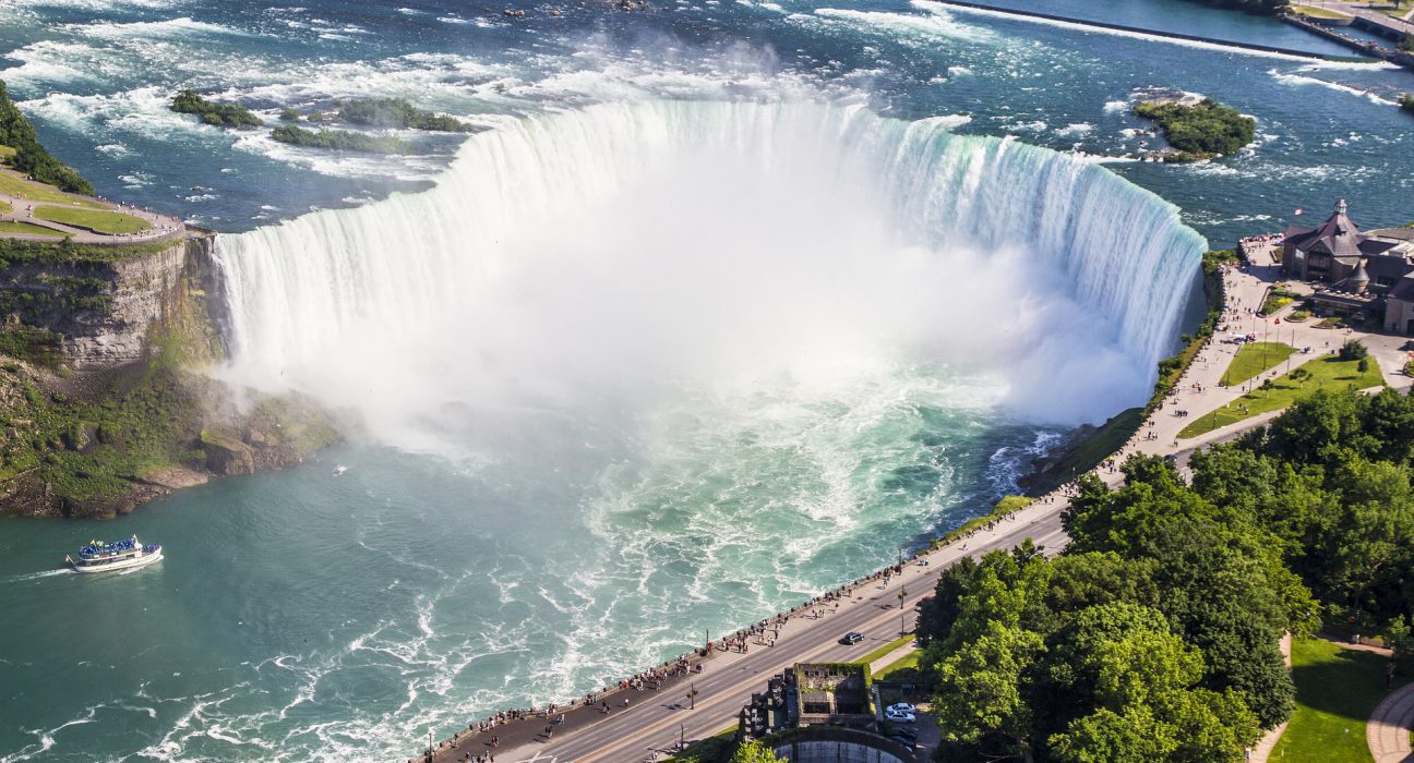 Aerial view of Niagara Falls with the large waterfall surrounded by lush greenery and a boat approaching the base of the falls. Tourists are visible near the edge.