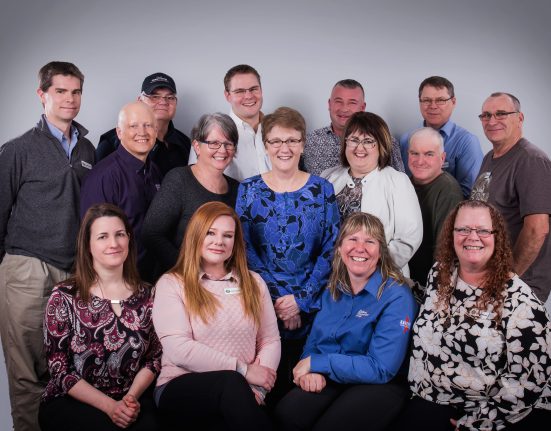 A group of 15 people of varying ages posing together against a gray background. They are dressed in a mix of casual and business attire.