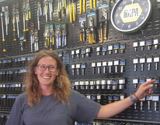 A person with glasses stands smiling in front of a display wall of tools and a clock, pointing to a section on the wall.