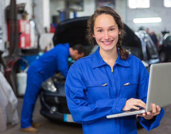 A person in blue coveralls holds a laptop, smiling, while another person works on a car engine in the background.