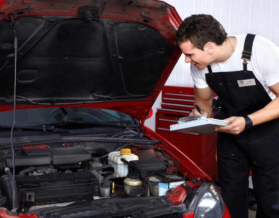 Mechanic in overalls inspects a cars engine with an open hood, holding a clipboard, in an auto repair shop.
