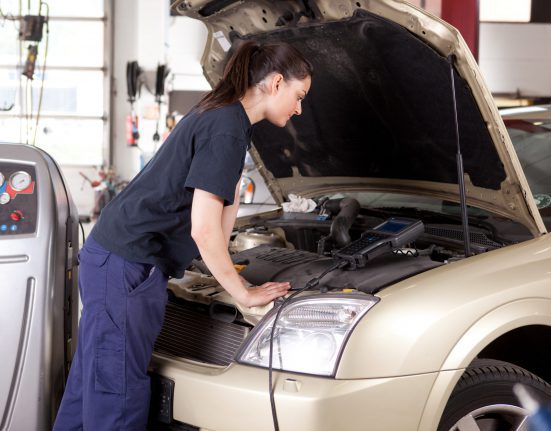 Mechanic inspecting the engine of a beige car in a garage setting, with diagnostic equipment nearby.
