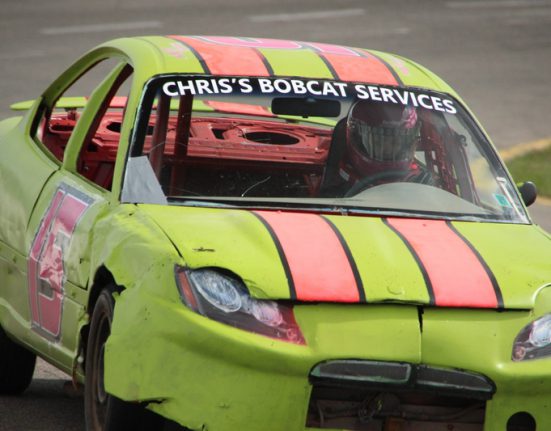 A bright green race car with pink stripes and Chriss Bobcat Services on the windshield races around a track. The driver wears a helmet.