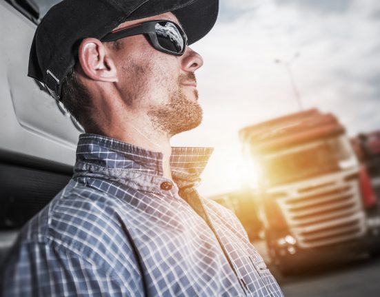 Man wearing a cap and sunglasses, leaning against a truck, with another truck in the background under a cloudy sky.