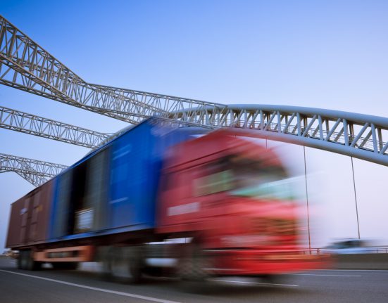A red cargo truck with a blue container speeds along a highway under a bridge, against a clear blue sky.