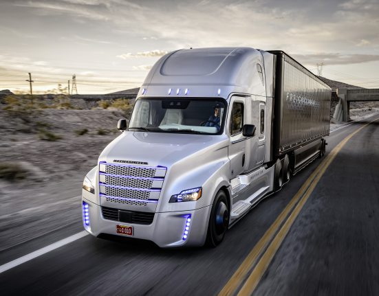 Silver semi-truck with modern design driving on a desert highway at sunset, with a bridge in the background.